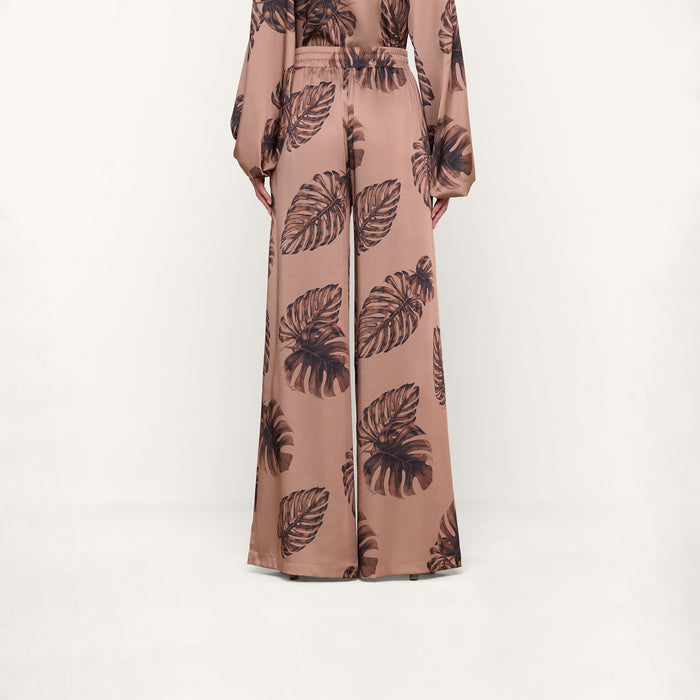 A person models the Lillian Satin Pant in an ultra-wide, high-waisted silhouette, featuring a large dark palm leaf print on a brown background. The matching long-sleeve top is partially visible. Only the lower body and hands are shown against a plain white backdrop.