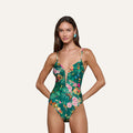 A woman stands against a plain light background, smiling with her hands on her hips, wearing the Lilou One-Piece Swimsuit featuring a green floral-butterfly print and a plunging V-wire neckline.