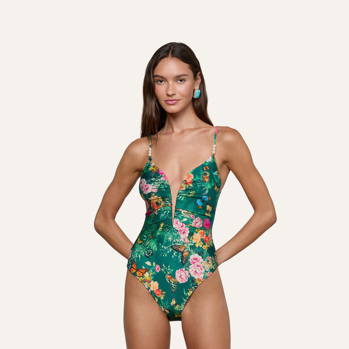 A woman stands against a plain light background, smiling with her hands on her hips, wearing the Lilou One-Piece Swimsuit featuring a green floral-butterfly print and a plunging V-wire neckline.