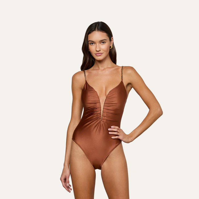 A woman models the Lilou One-Piece Swimsuit—a shiny brown swimsuit featuring a deep v-wire plunge neckline, thin straps, and a high-leg cut—as she stands with one hand on her hip against a plain light background.