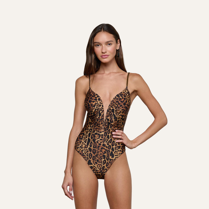 A woman with long brown hair stands with one hand on her hip, wearing the Lilou One-Piece Swimsuit featuring a plunging neckline and leopard print. She has a neutral expression and is posed against a plain light background.