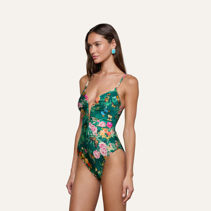 Model wears the Lilou One-Piece Swimsuit, featuring a green base with a vibrant floral-butterfly print, a plunging V-wire neckline, and thin shoulder straps, standing against a plain light background.