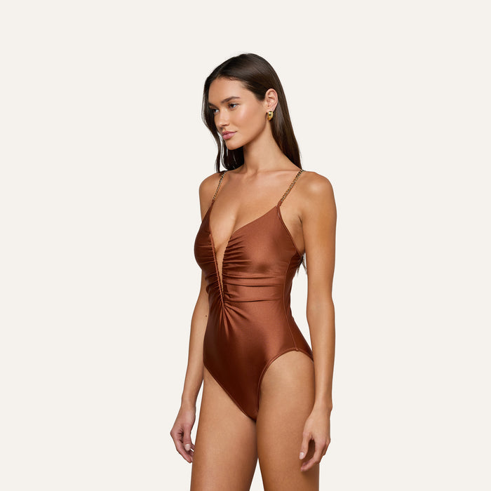 A woman stands against a plain light background, wearing the Lilou One-Piece Swimsuit in a shiny, deep brown shade. This swimsuit features a plunging v-wire neckline, thin straps, and a high-leg cut that accentuates her silhouette.