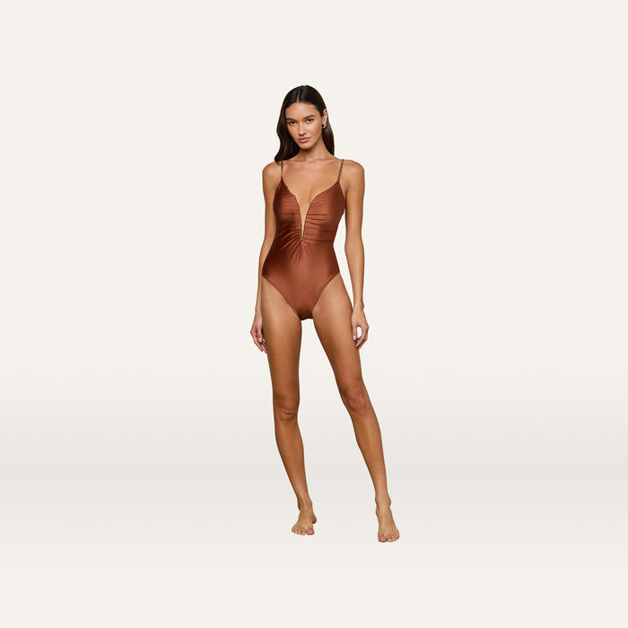 A woman stands against a plain white background, wearing the Lilou One-Piece Swimsuit—a shiny brown swimsuit featuring thin straps, a high-leg cut, and a v-wire plunge neckline—looking at the camera.