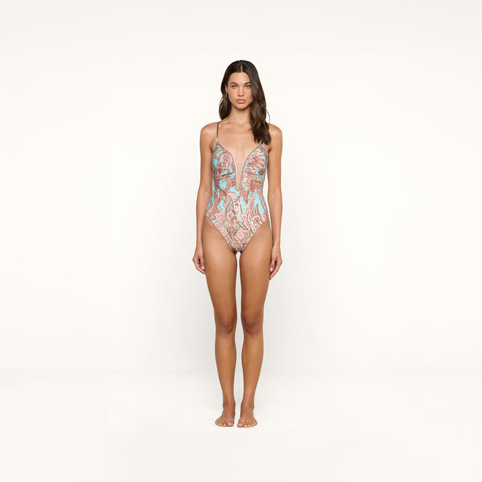 A woman stands facing forward against a plain white background, wearing the Lilou One-Piece Swimsuit, which features a pastel pattern and brass-chain straps.