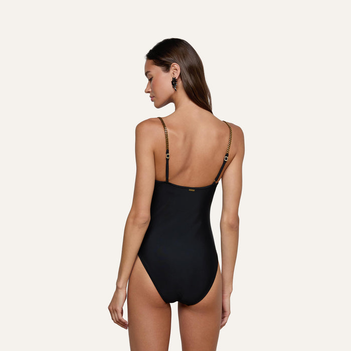 A woman with long brown hair stands with her back to the camera against a plain light background, wearing the Lilou One-Piece Swimsuit—a black v-wire plunge style featuring thin brass-chain straps.