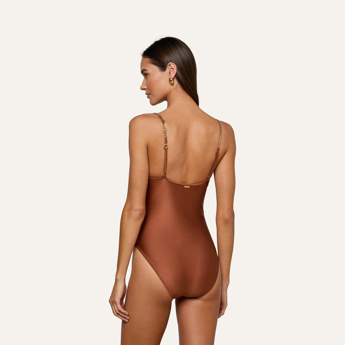 Wearing the Lilou One-Piece Swimsuit—a metallic brown design with a high-leg cut and thin, chain-like straps—a woman with long brown hair stands against a plain light background, facing away and looking over her shoulder.