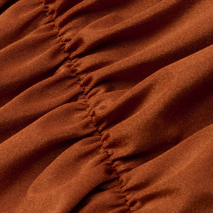 A close-up of the Lilou One-Piece Swimsuit in rust shows gathered fabric with visible vertical stitching that creates soft folds and textured, ruched lines. The material looks smooth and slightly shiny, highlighting the high-leg cut design.