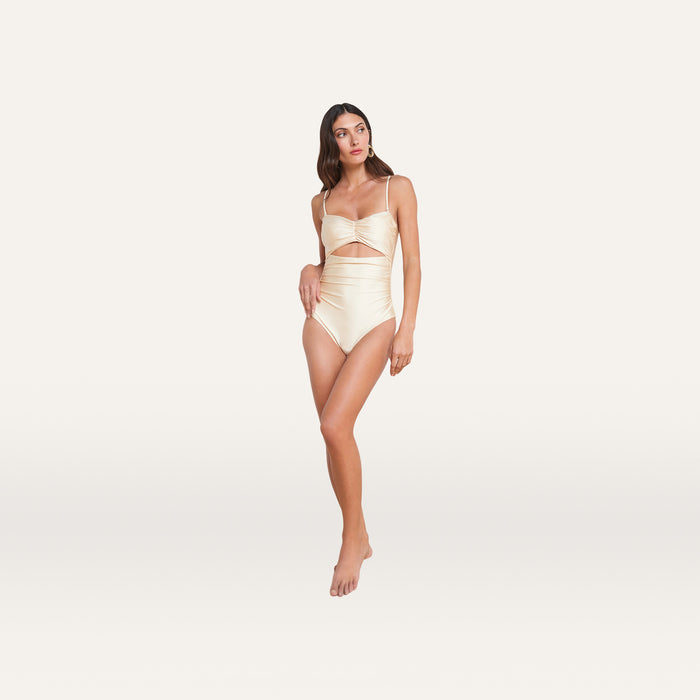 A woman stands barefoot on a light background, wearing the Lily Cut-Out One-Piece Swimsuit in a shiny, light beige hue. The swimsuit features cut-out details at the waist, thin straps, and a softly ruched bodice. She looks slightly to her left with a neutral expression.