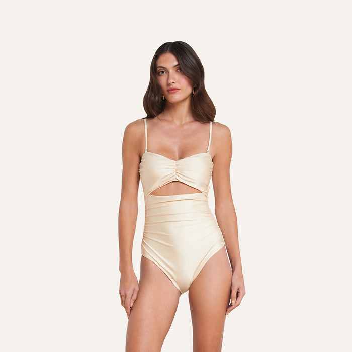 A woman wearing the Lily Cut-Out One-Piece Swimsuit—a light beige, high-waisted one-piece featuring a sweetheart neckline, ruched bodice, thin straps, and a cutout below the bust—stands against a plain off-white background.