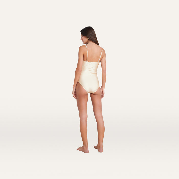A woman with long brown hair stands barefoot, facing away, wearing the Lily Cut-Out One-Piece Swimsuit in cream, featuring a ruched bodice and thin straps, set against a plain white background.