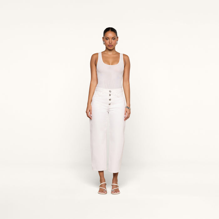 A woman stands facing forward against a plain white background, wearing a sleeveless white top, the Liora Cropped Jean—high-rise, white wide-leg pants with buttons—white sandals, and hoop earrings.