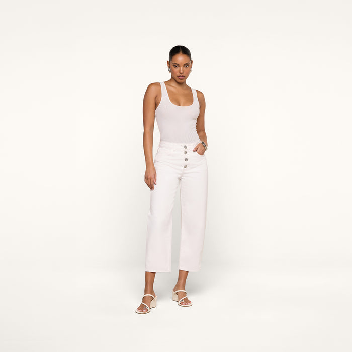 A woman stands against a plain white background wearing a sleeveless beige top, the Liora Cropped Jean—high-rise, wide-leg white pants with button details—and white sandals. Her hands are in her pockets and she has a neutral expression.