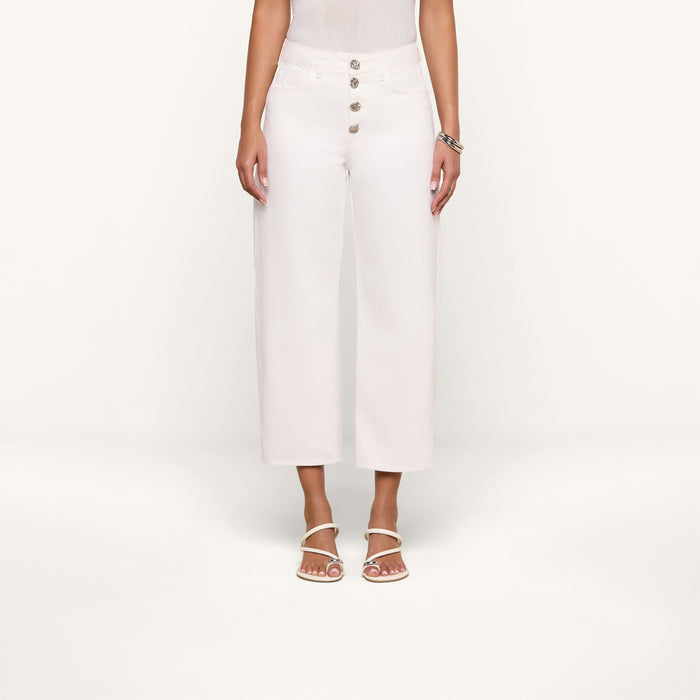 A woman wearing the Liora Cropped Jean—white high-rise, wide-leg jeans with large buttons—paired with a white sleeveless top, white heeled sandals, and a bracelet, stands against a plain white background. Her face is out of frame.