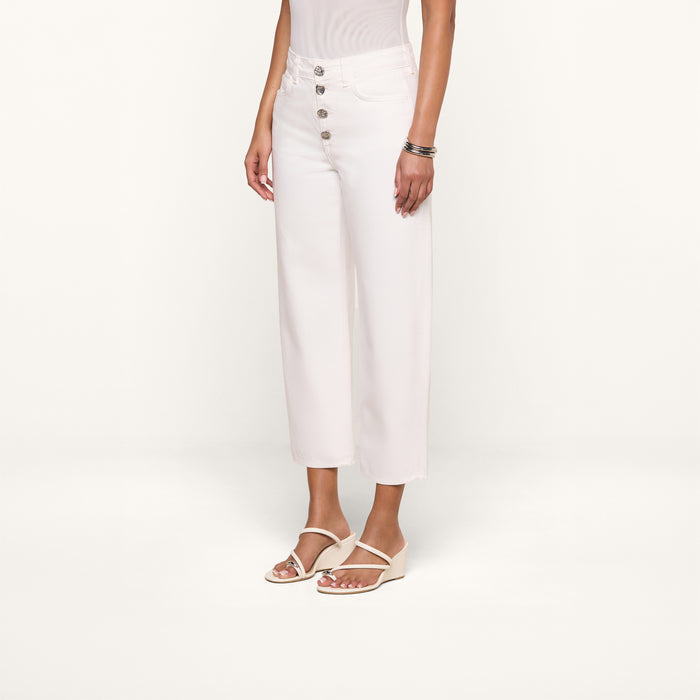 Wearing the Liora Cropped Jean—a pair of high-rise, wide-leg white jeans with large silver buttons—a person stands against a plain white background, styled with a sleeveless white top, white sandals, and silver bracelets.