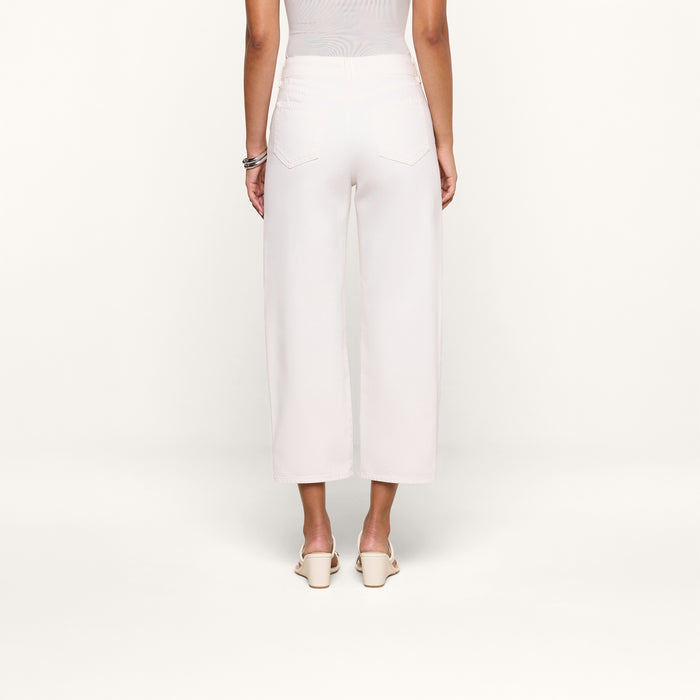 A person stands facing away from the camera, wearing the Liora Cropped Jean in white—a high rise, wide-leg style—paired with a sleeveless top, white heeled shoes, and bracelets, against a plain white background.