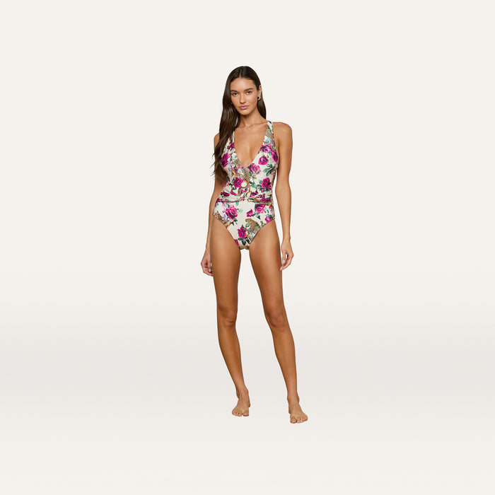 A woman stands on a plain background, wearing the Lisa Plunge One-Piece Swimsuit, which features a rose leopard print and pink floral pattern. She is barefoot, facing forward, and smiling slightly.