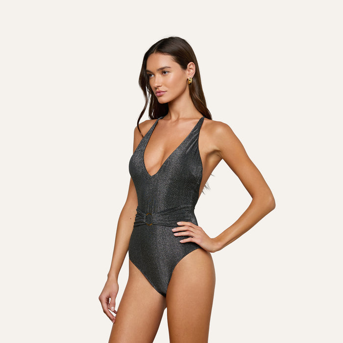 A woman poses against a plain, light background wearing the Lisa Plunge One-Piece Swimsuit, featuring a metallic finish, deep plunge neckline, and a belted waist detail.