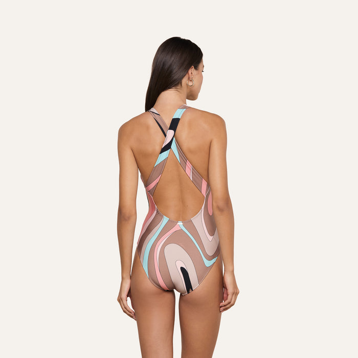 A woman with long brown hair models the Lisa Plunge One-Piece Swimsuit, which features an abstract swirl print in pink, white, blue, and black. The swimsuit has a racerback and an open back design. She is standing and facing away from the camera.