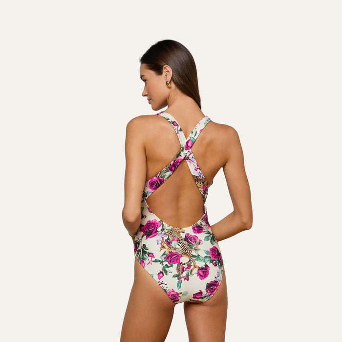 A woman faces away, wearing the Lisa Plunge One-Piece Swimsuit, which features a deep plunge neckline, pink rose and bird prints, crisscross straps, and an open back. The UPF 50 swimwear stands out against the plain, light-colored background.