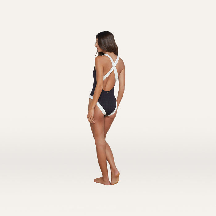 A woman stands barefoot against a plain white background, looking to the side, wearing the Lisa Plunge One-Piece Swimsuit in black. The swimsuit features white cross-back straps and offers moderate bottom coverage.