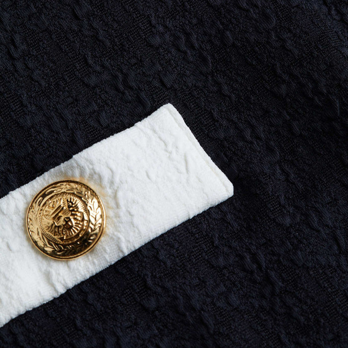 A close-up of the Lisa Plunge One-Piece Swimsuit showcases textured black fabric with a white strip and a gold decorative button featuring an embossed design, evoking the look of bouclé.