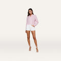 A woman with long hair and a relaxed expression stands against a plain light background, wearing the high-rise white Lissania Jean Short with a light pink long-sleeve button-up shirt and white heeled sandals.