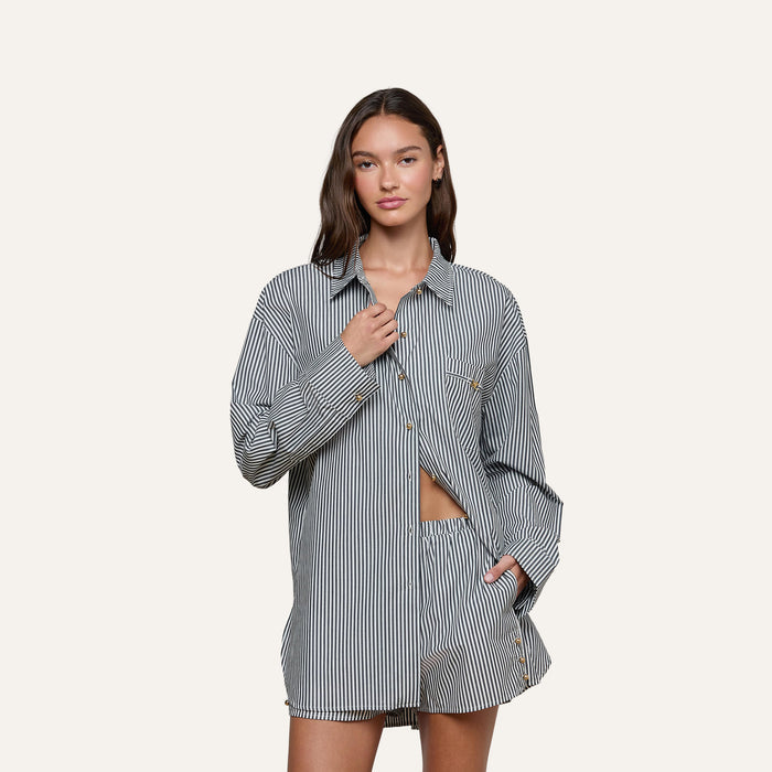 A woman stands against a plain background wearing the Lorena Cover-Up Shirt, a long-sleeved black-and-white striped resort top with gold buttons, paired with matching shorts. She has one hand at her collar and the other in her pocket, looking confidently at the camera.