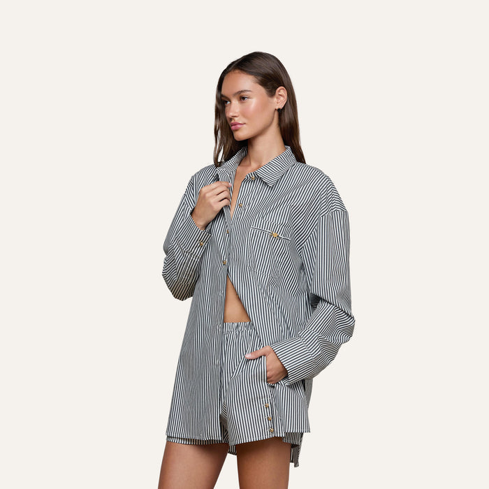 A woman wearing the Lorena Cover-Up Shirt, a loose, long-sleeve resort top in black-and-white striped cotton poplin with matching shorts, stands against a plain light background—one hand holding the shirt collar and the other tucked into a pocket.