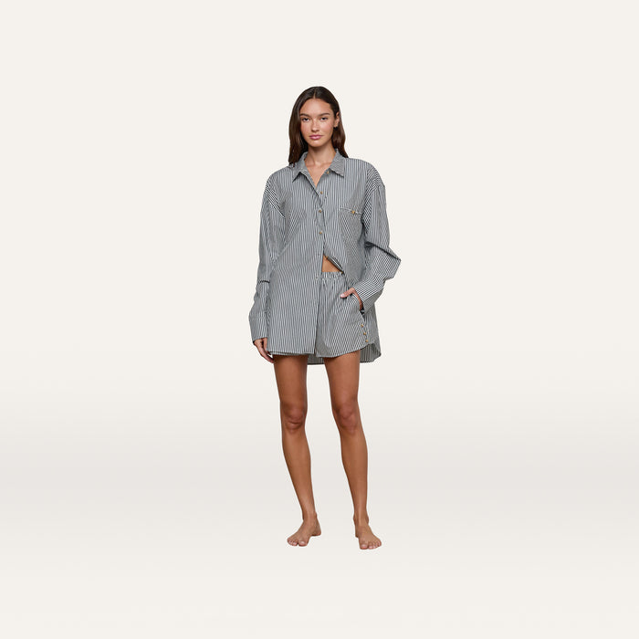 A woman stands barefoot against a plain white background, wearing the loose-fitting Lorena Cover-Up Shirt with gold buttons and matching striped shorts.