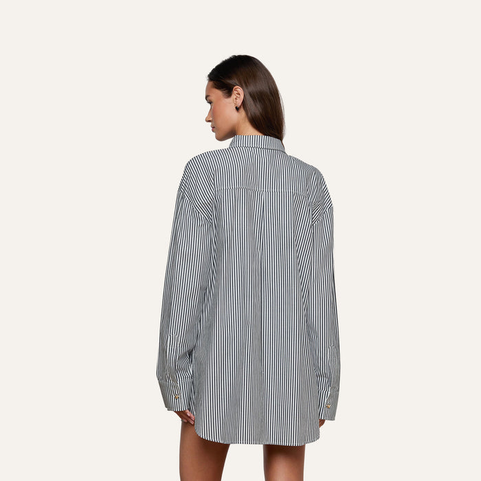 A woman with straight brown hair is standing with her back to the camera against a plain, light background, wearing the Lorena Cover-Up Shirt—a long-sleeve, black-and-white striped resort style.