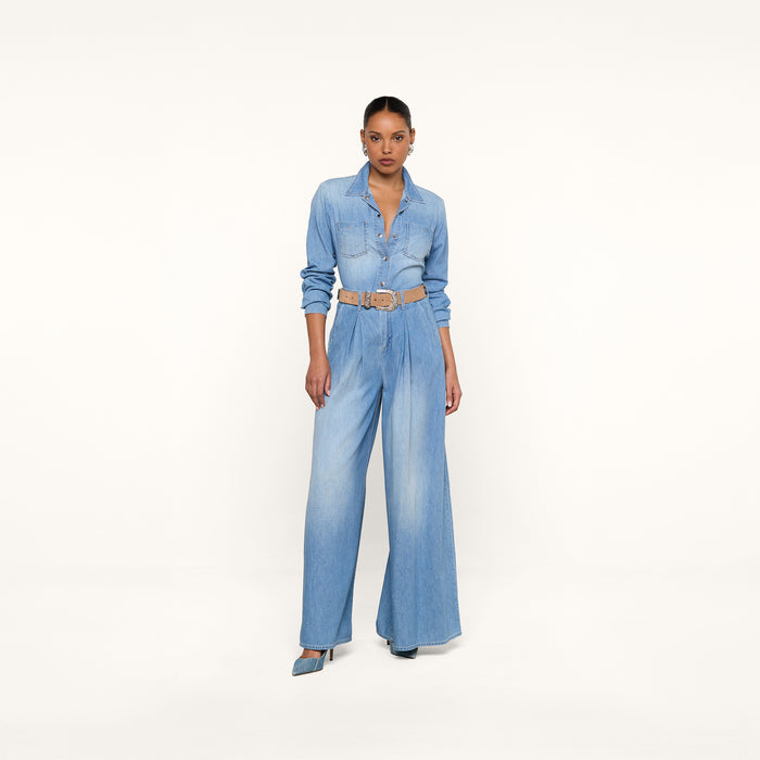 A woman stands against a white background wearing the Lorenza Palazzo Jean, a light blue featherweight chambray denim jumpsuit with a stylish high-rise palazzo leg silhouette, paired with a tan belt and blue heels, looking confidently at the camera.