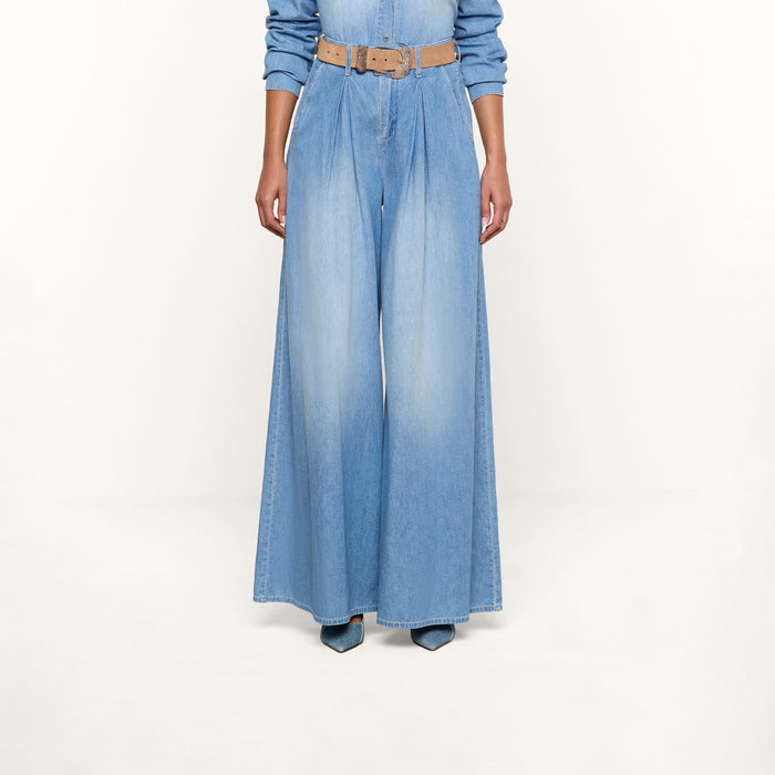 Wearing the Lorenza Palazzo Jean in light blue denim, paired with a tan belt and pointed-toe blue shoes, a person is shown from the shoulders down against a plain white background.