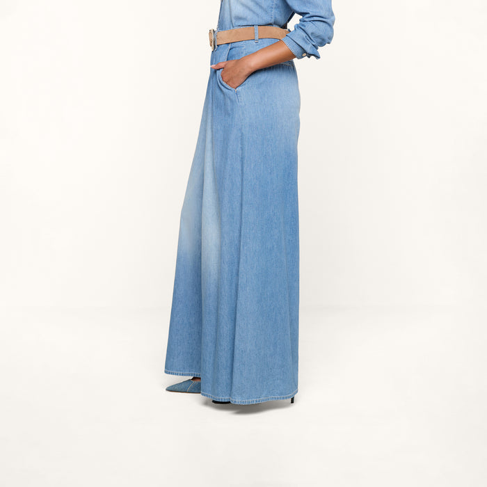 A person is wearing the Lorenza Palazzo Jean in light blue featherweight chambray denim, styled with a wide tan belt, hands in pockets, and a matching denim shirt. The background is plain white.