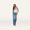 A woman stands against a plain, light background with her hands in her pockets, wearing a fitted white tank top and the Louane Wide-Leg Flare Jean in stretch denim.