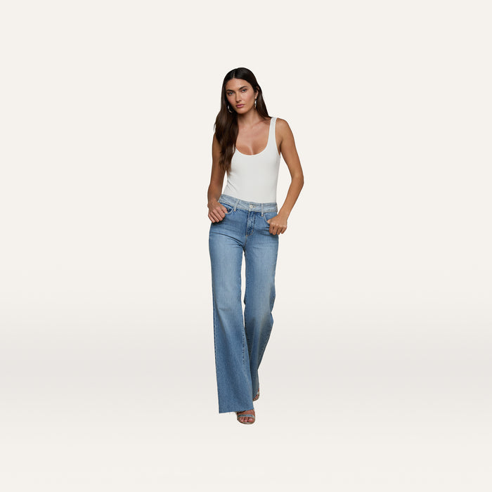 A woman stands against a plain, light background with her hands in her pockets, wearing a fitted white tank top and the Louane Wide-Leg Flare Jean in stretch denim.