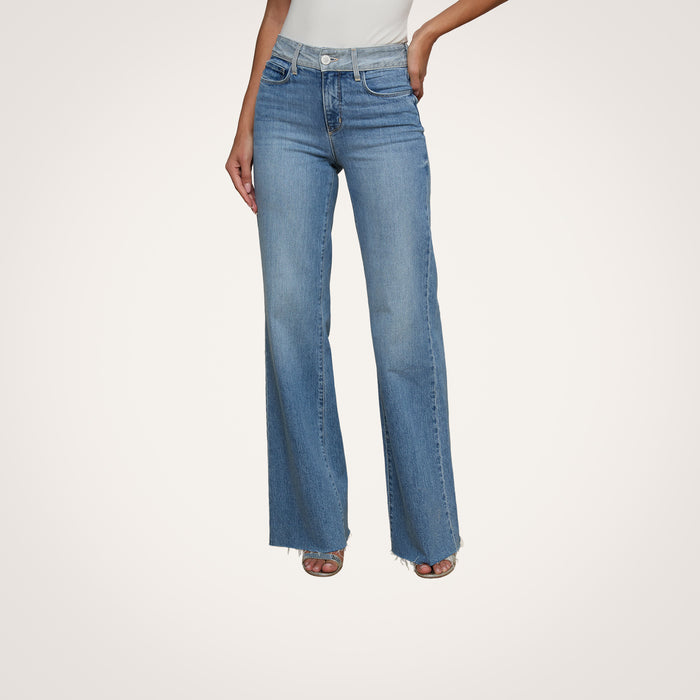 A woman models the Louane Wide-Leg Flare Jean in light blue, featuring a high-rise fit and frayed hems, paired with a white sleeveless top. She stands against a plain light background with her hands at her sides and her face out of view.