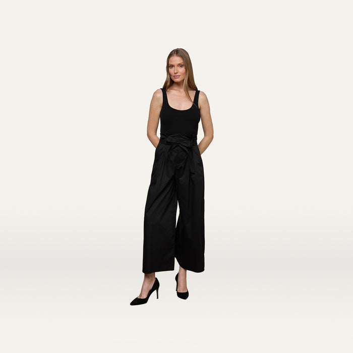 A woman stands against a plain white background wearing a sleeveless black top, black heels, and the Lowen Pant—wide leg black poplin pants featuring a waist tie.