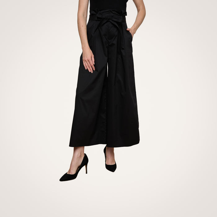 A person wearing the Lowen Pant—a pair of high rise black poplin pants with a waist tie—paired with black high heels, standing against a plain light background.