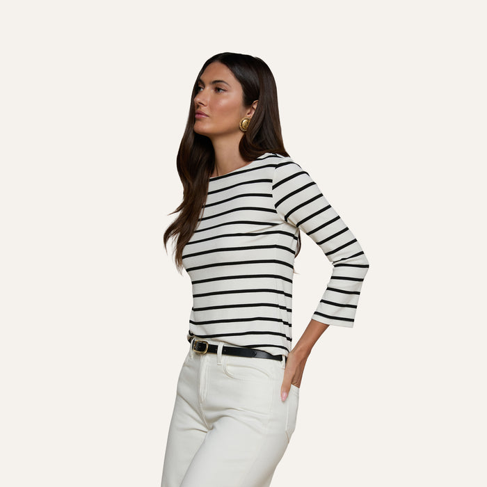 Wearing the Lucille Striped Boatneck Top—a black and white, long-sleeve shirt featuring a boat neckline—paired with white pants and a black belt, the woman stands with one hand in her pocket against a plain background. The timeless stripes of the top add classic appeal to her look.