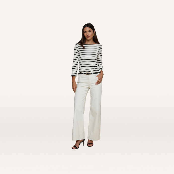 A woman stands against a plain background wearing the Lucille Striped Boatneck Top, paired with white wide-leg pants, a black belt, and black high-heeled sandals. With one hand in her pocket and a neutral expression, she captures the effortlessly chic appeal of timeless stripes.