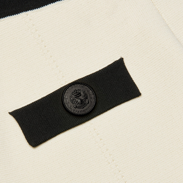 Close-up of the cream-colored Lumi Knit Micro Short’s power knit fabric, featuring a black rectangular patch adorned with a round, embossed black button with text and a crest-like emblem in the center.