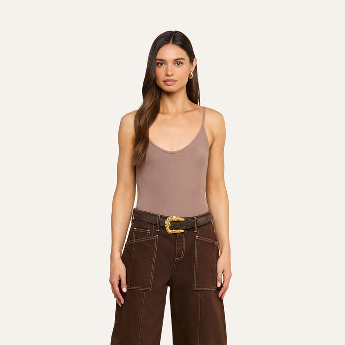 A woman with long brown hair wears a Lunara Camisole crafted from soft micro modal ribbed fabric, paired with dark brown pants featuring white stitching and a decorative belt, standing against a plain light background.