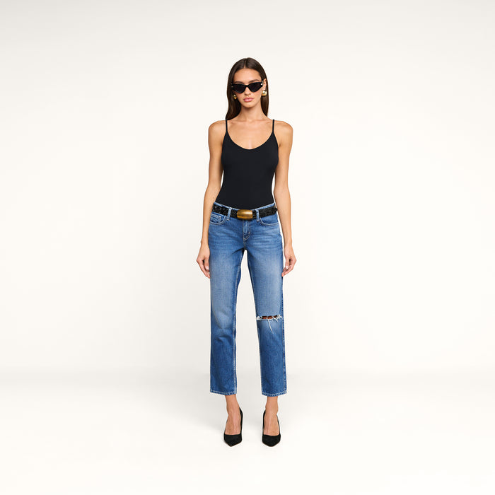 Woman wearing a black Lunara Camisole made from soft micro modal ribbed fabric, blue ripped jeans with a belt, black high heels, and sunglasses, standing against a plain white background.