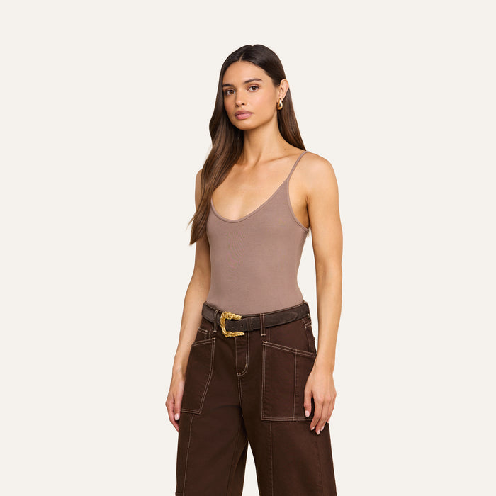 A woman with long brown hair wears a taupe Lunara Camisole made from soft micro modal ribbed fabric, paired with high-waisted brown belted pants, standing against a plain light background.