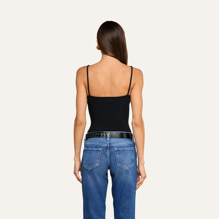 A woman stands facing away, wearing the Lunara Camisole—a black ribbed fabric spaghetti strap top—paired with blue jeans and a black belt. Her long brown hair is straight and loose, and her arms rest relaxed at her sides against a plain light background.