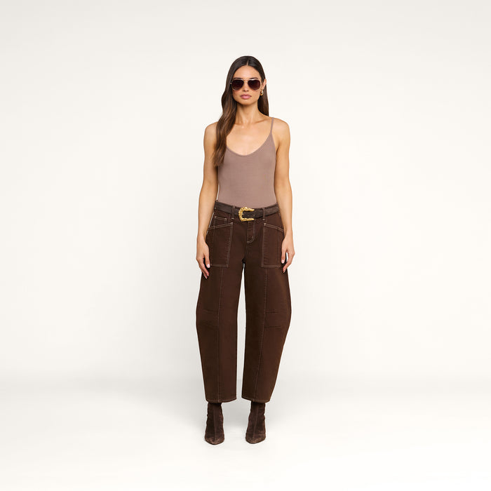 A woman wearing sunglasses, a taupe Lunara Camisole crafted from micro modal ribbed fabric, brown high-waisted belted pants, and brown boots stands against a plain white background.