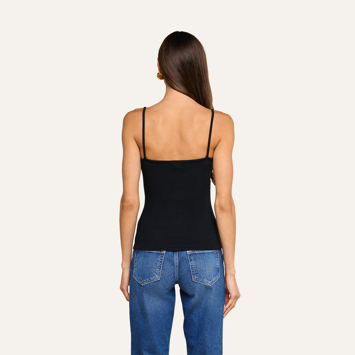 A woman stands with her back to the camera, wearing the Lunara Camisole in black ribbed fabric and blue jeans against a plain, light background.