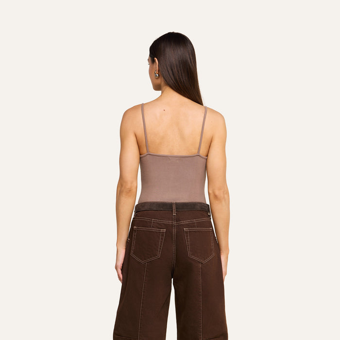 A woman with long brown hair, wearing a taupe Lunara Camisole crafted from ribbed micro modal fabric and dark brown wide-leg pants, stands facing away from the camera against a plain light background.