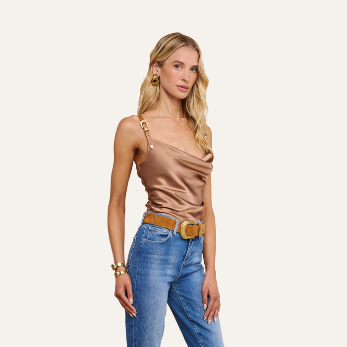 A woman wearing the Lux Silk Buckle-Strap Camisole—a shiny bronze cowl-neck top with vegan leather straps—paired with blue jeans, a tan belt, gold earrings, and a bracelet, stands against a plain white background.